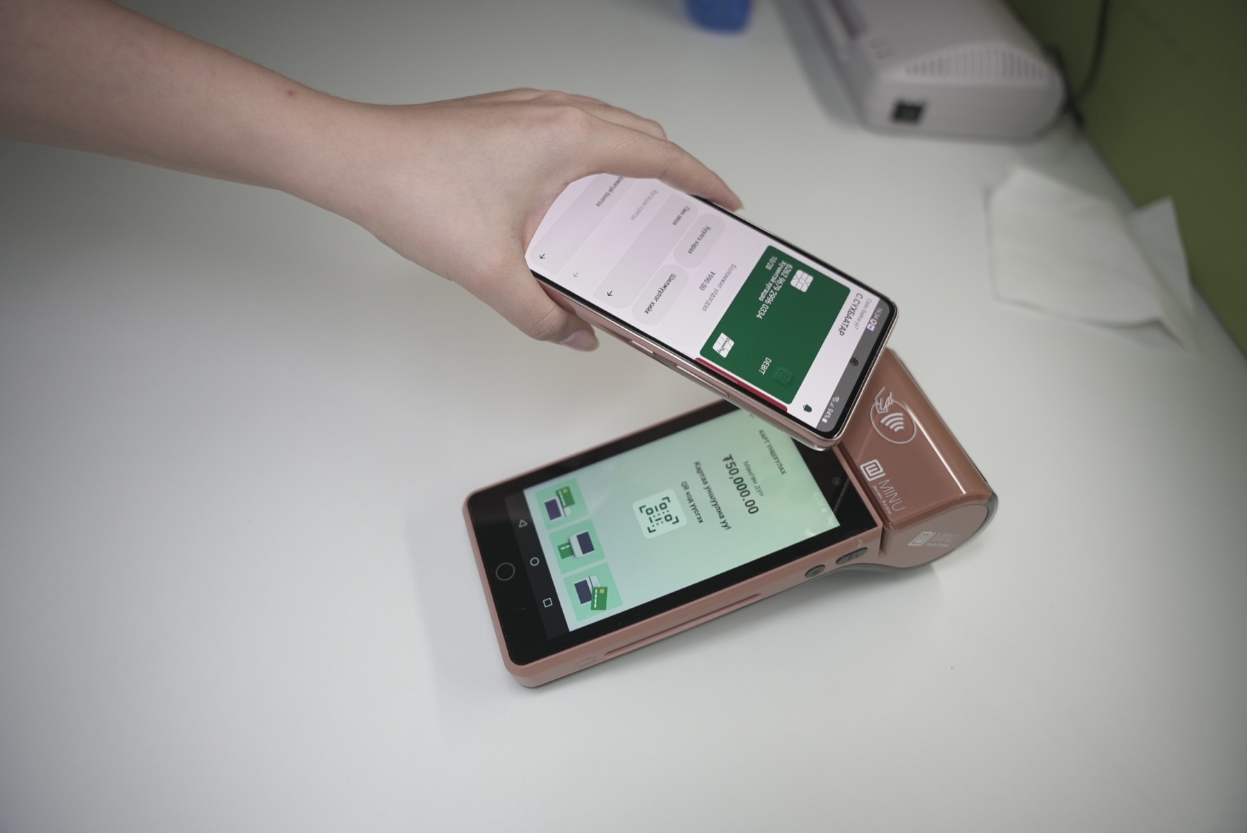 A hand holds a phone showing a green bank card on screen and taps it against a rose gold Minu POS terminal lying on a white desk. The terminal's screen shows a QR code and an amount.